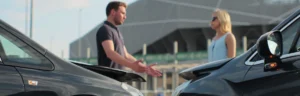 A man in a dark shirt and a blonde woman in a light blue dress are discussing accident details between two damaged vehicles, seeking auto accident legal help.