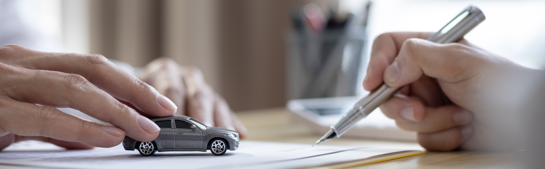 A person's hands are positioning a gray toy sedan on papers while holding a silver pen representing an auto accident settlement calculator and claim documentation process.