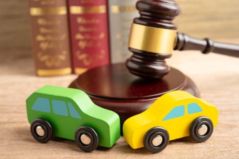 A green SUV and yellow sedan toy cars are positioned on a wooden desk in front of a judge's gavel and law books for a car accident attorney consultation.
