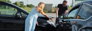 A blonde woman in a light blue dress and a man in a black shirt are inspecting front-end damage on a black car, discussing car accident compensation options.