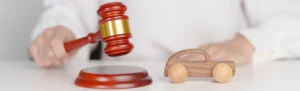 A person's hand holding a wooden gavel with a gold band above a toy car and sound block representing a local attorney handling a vehicle accident case.