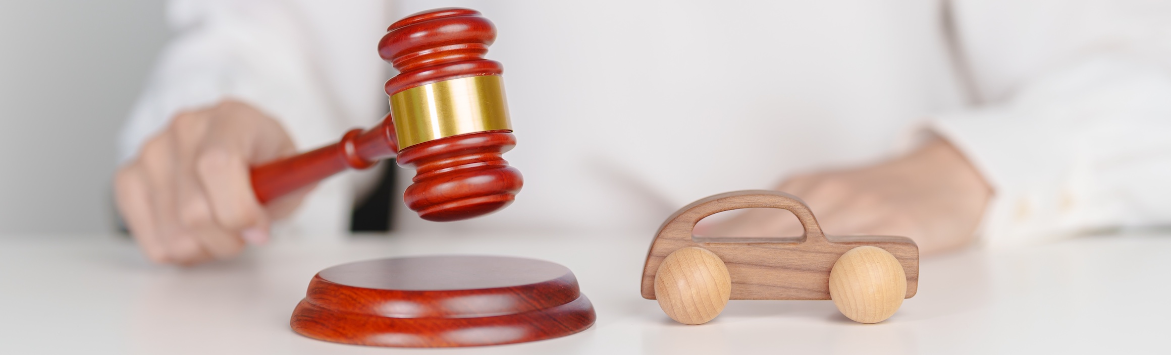 A person's hand holding a wooden gavel with a gold band above a toy car and sound block representing a local attorney handling a vehicle accident case.