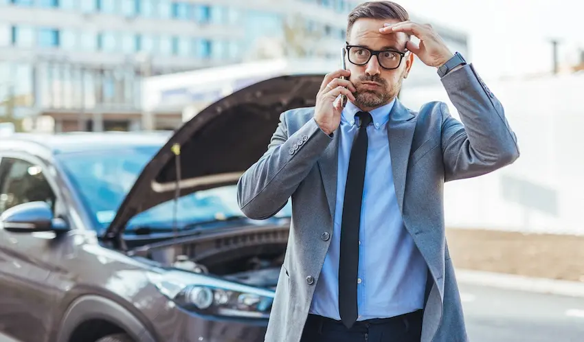 A stressed businessman is on the phone standing near his broken-down vehicle following a car accident requiring roadside assistance in Tennessee.