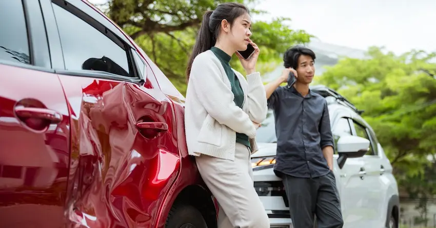 A woman is calling for help after a car accident in Tennessee and is leaning against her damaged red car while the other car accident victim who is standing in front of his white car is also on his phone.