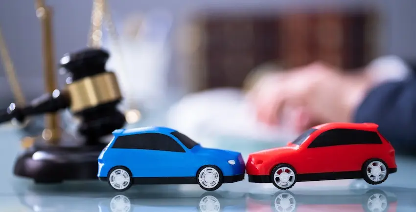 A blue and red toy car staged in a front-end collision sit on a glass desk in front of a judge's gavel and scales of justice illustrating how different car accident types such as head-on carry distinct legal implications that an attorney from Mogy Law Firm can help victims navigate.