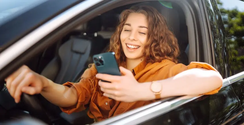 A young woman with curly hair smiles and looks down at her smartphone while seated behind the wheel of a car with the window down illustrating the distracted driving behavior that is one of the leading causes behind multiple car accident types on roads today in Tennessee.