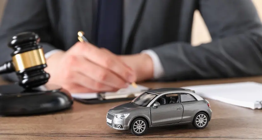 A suited attorney from Mogy Law Firm writes notes at a desk beside a black gavel with a silver model car in the foreground representing a rollover accident attorney reviewing case details and building a legal strategy on behalf of an injured client.