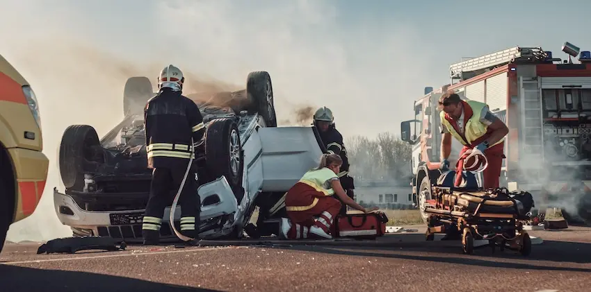 Firefighters and paramedics with a stretcher work urgently to rescue a victim from a smoking overturned vehicle on a road with emergency trucks in the background illustrating the life-threatening nature of the crashes that prompt injured survivors to contact a rollover accident attorney in Tennessee.
