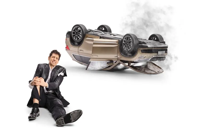 A injured man in a suit sits on the ground wincing in pain beside a smoking overturned vehicle representing a rollover accident victim who needs to contact a rollover accident attorney from Mogy Law Firm in Tennessee to recover compensation for his injuries and damages.