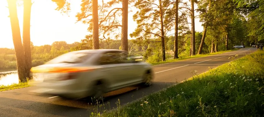 A white sedan speeds along a narrow tree-lined road at sunset with motion blur emphasizing its high velocity representing the role that excessive speeding plays in some of the most dangerous car accident types including head-on collisions and roadway departures in Tennessee.
