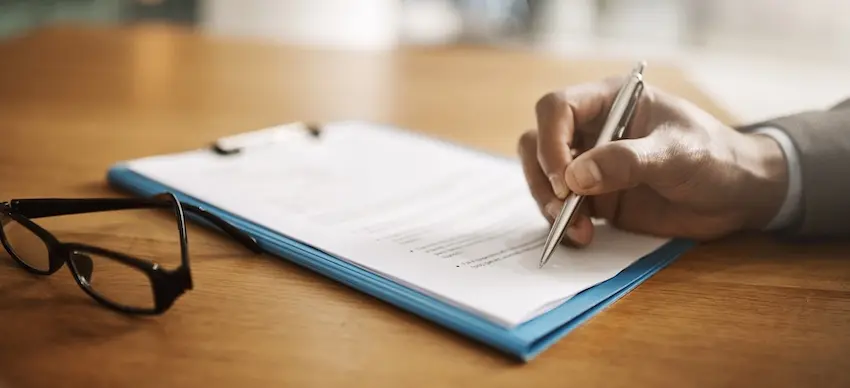 A hand in a gray sleeve holding a silver pen signs a document secured to a blue clipboard on a wooden desk with a pair of black-framed glasses resting beside it- representing the formal signing process that finalizes a settlement offer and brings resolution to an accident victim's compensation claim in Tennessee.