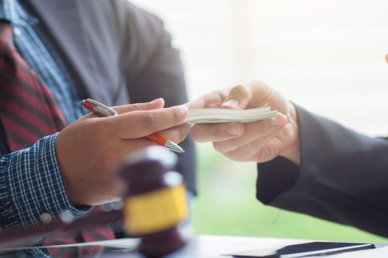A man in a blue plaid shirt and red striped tie holds a pen while accepting a stack of cash handed across a desk with a wooden judge's gavel and legal documents visible in the foreground illustrating the exchange of compensation when an injury victim successfully negotiates and accepts a fair settlement offer through their attorney from Mogy Law Firm in Tennessee.