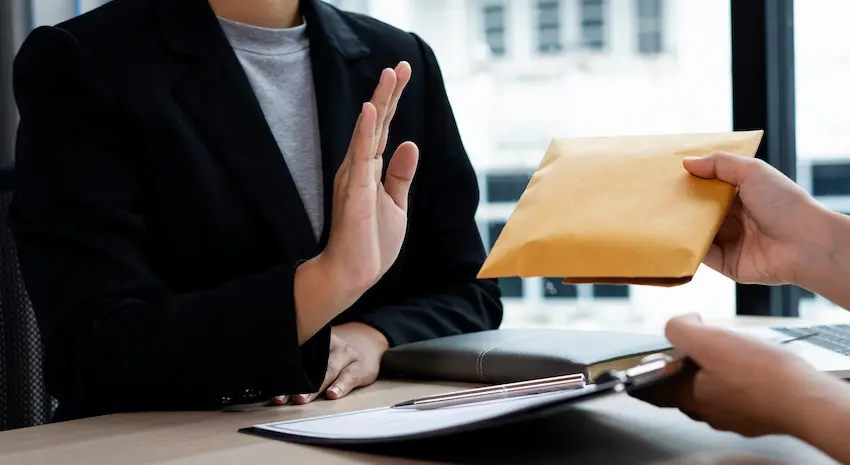 A person in a black blazer and gray shirt raises one hand firmly in refusal as another person extends a brown envelope across a desk with a clipboard- notepad- and pen visible on the surface- depicting an accident victim declining an inadequate settlement offer from an insurance company in pursuit of full and fair compensation in Tennessee.