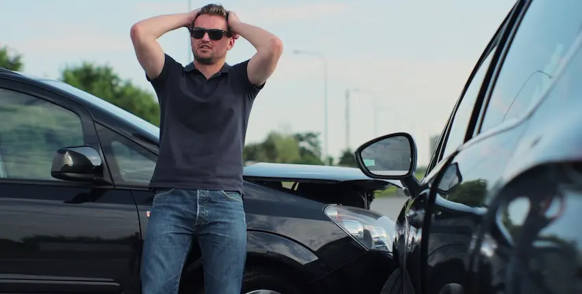 A frustrated man wearing dark sunglasses and a navy polo shirt stands between two black vehicles that have collided front to back- both hands raised and clasped behind his head in a gesture of shock and disbelief with the crumpled hoods of both cars visible on either side of him and a blurred roadway with utility poles and green trees stretching across the background — capturing the stressful moment immediately after a crash when a driver must begin thinking about filing an insurance claim with help from Mogy Law Firm in Tennessee.