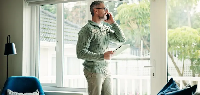 A middle-aged man with short gray hair- a neatly trimmed beard- and glasses stands near a large floor-to-ceiling window in a bright- modern living room- holding a white tablet in one hand while pressing a smartphone to his ear with the other- wearing a sage green cable-knit zip-up sweater and khaki pants- with a blue accent chair- a dark floor lamp- and lush green trees visible through the glass behind him — depicting an accident victim making a call to initiate an insurance claim from the comfort of his home in Tennessee.