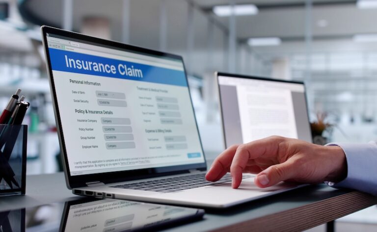 A person's hand in a dark business blazer sleeve reaches across a reflective black desk to type on a silver laptop displaying a blue-headed online form titled "Insurance Claim" with visible fields for personal information including date of birth and social security number as well as sections for policy and insurance details such as policy number- group number- and employer name- with a second open laptop and a small potted plant blurred in the background of a bright modern office — showing the digital process of completing and submitting an insurance claim with help from Mogy Law Firm.