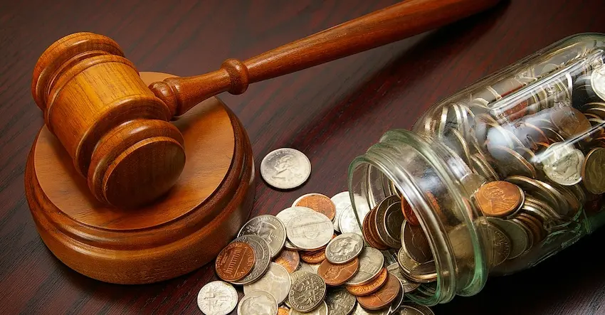 A polished honey-toned wooden judge's gavel rests on its circular sound block on the left side of a dark mahogany surface while a clear glass mason jar tipped on its side spills a large mixed collection of coins — including quarters- dimes- nickels- and pennies — across the table to the right with the scattered coins reflecting the financial compensation at stake when pursuing a legal case tied to an insurance claim in Tennessee.