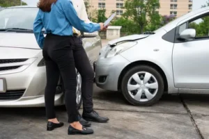Two drivers standing outside after a car accident, one holding a clipboard near a damaged front end bumper in a parking lot.