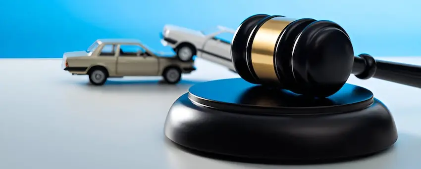 A sleek black judge's gavel with a wide gold band rests upright on its matching black circular sound block in the sharp right foreground while two vintage-style cream-colored toy sedans are staged in a rear-end collision in the softly blurred left background- one car riding up over the back of the other all set against a vivid solid blue backdrop on a white surface — a clean legal concept image representing the courtroom proceedings and attorney involvement from Mogy Law Firm-required to recover compensation for medical expenses and lost wages following a car accident claim.