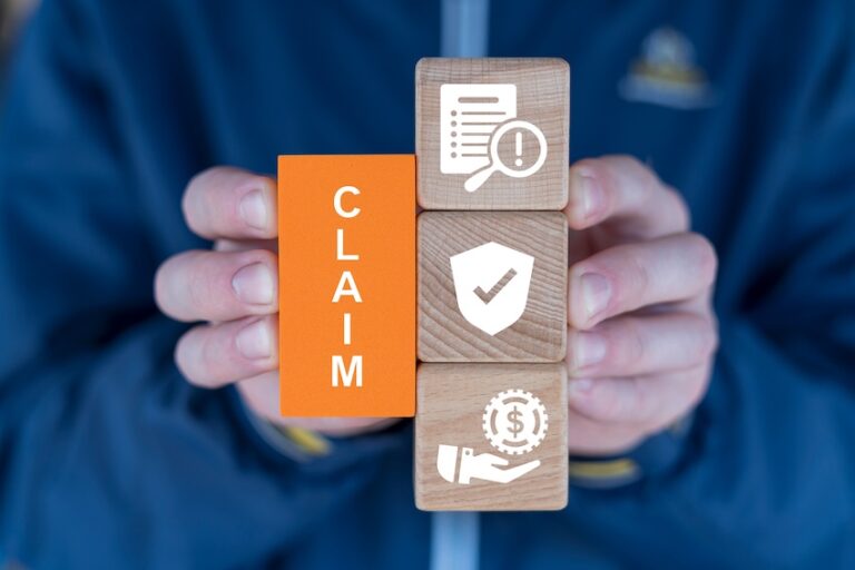 A lawyer from Mogy Law Firm in Tennessee is wearing a dark blue jacket holds both hands around a vertical stack of three natural wood square blocks the left side of the stack featuring a flat orange rectangular tile printed with the word CLAIM in bold white vertical capital letters while the three wooden cube faces visible on the right display white illustrated icons representing the claims process — the top cube showing a document under a magnifying glass with an exclamation point the middle cube showing a shield with a checkmark symbolizing coverage approval and the bottom cube showing a hand holding a coin gear symbol representing financial compensation — a tactile visual representation of the structured steps involved in filing a claim to recover lost wages and injury-related expenses.